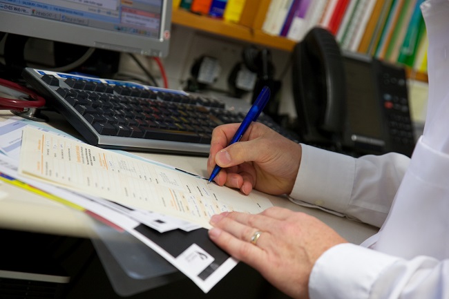 Close-up of the hands of a White male GP completing a form at his desk